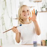 Happy and playful cute freckled blonde girl dressed in white, holding brush in one hand and showing another hand, which she messed up with paint. Children and happiness concept. Isolated shot, horizontal